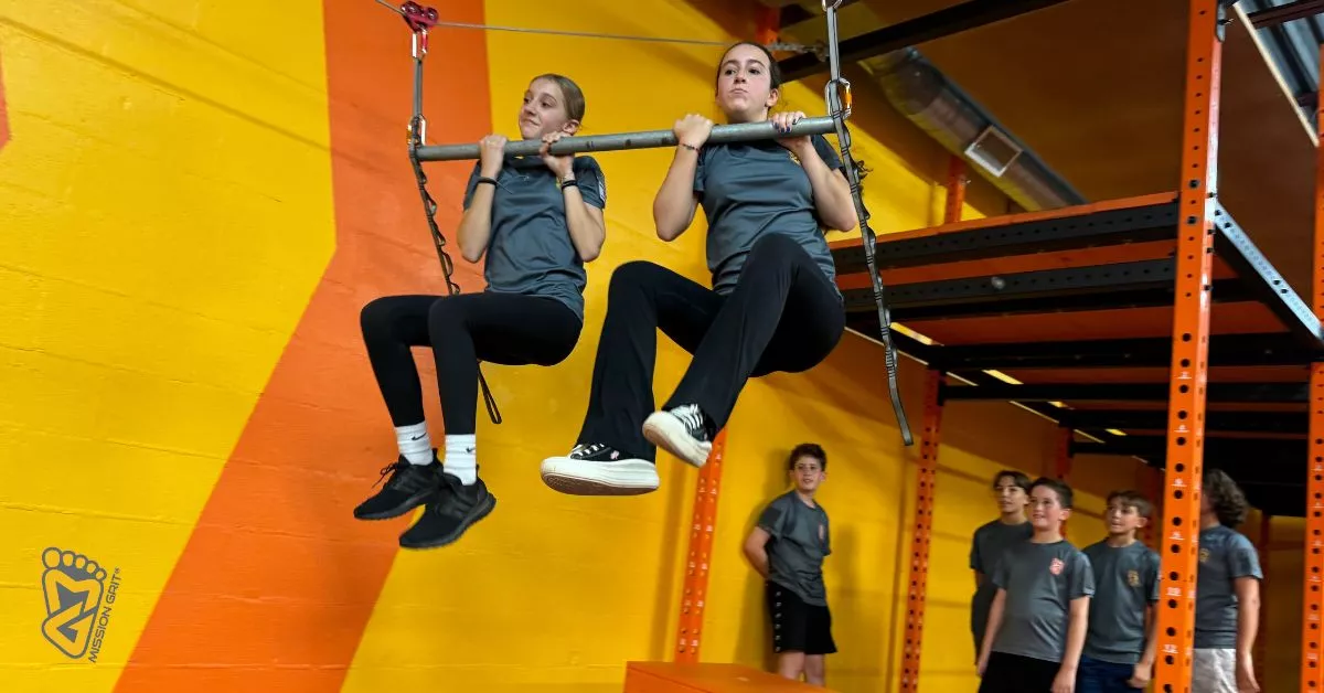 Two girls practice hanging bar obstacles during ninja warrior training for kids inside Mission Grit’s brightly colored gym, building strength and confidence as classmates watch.