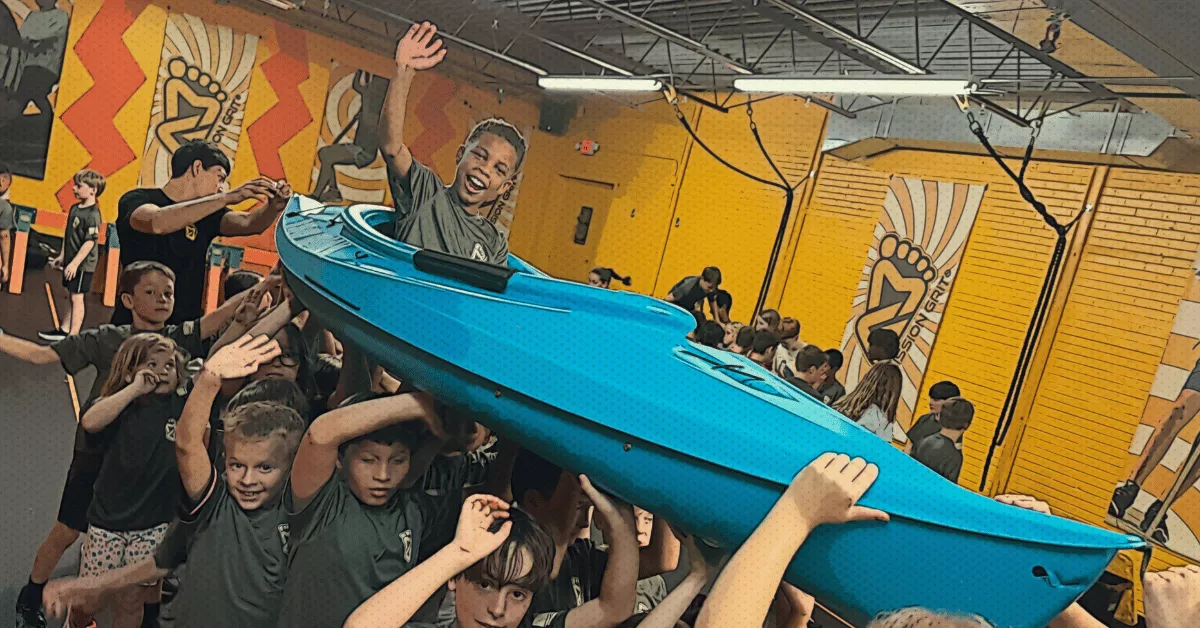 Children lifting a kayak together indoors during a cooperative team-building activity at spring camp.