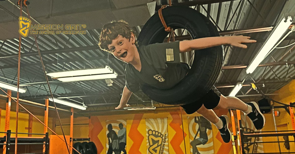 Boy smiles while swinging through an indoor obstacle course on a suspended tire at Mission Grit.