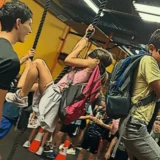 Children wearing harnesses swing on ropes through an indoor obstacle course while a coach guides them and other campers watch.