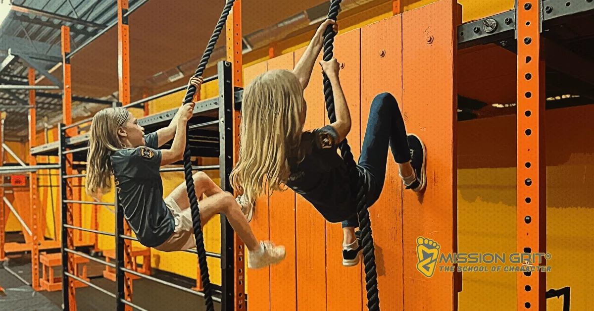 Two girls climb ropes on an indoor obstacle wall at Mission Grit during a structured physical challenge.