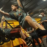 Kids at Mission Grit's Middle School Preparation Charlotte & Fort Mill swing across thick ropes in an indoor obstacle gym, while other students watch in the background.