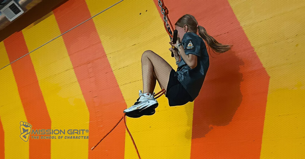 A girl in athletic clothes rides a zipline-style obstacle across an indoor gym with orange and yellow walls and Mission Grit branding.