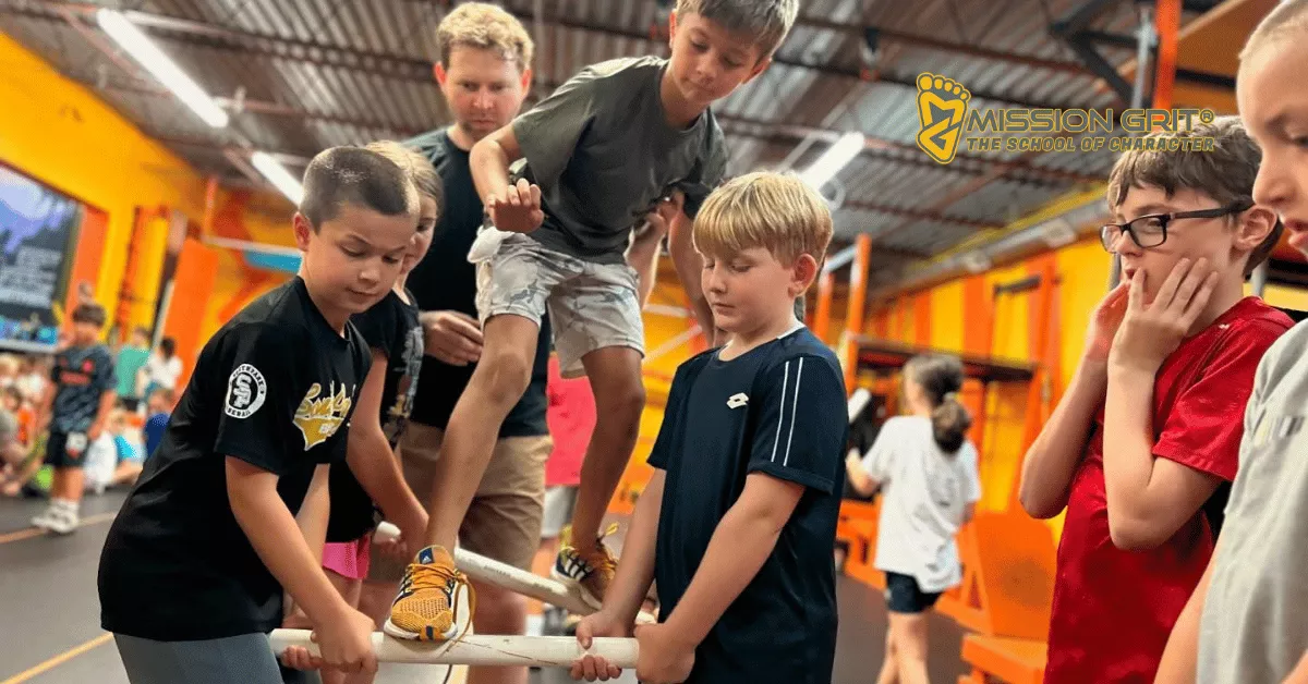 Kids work together to balance a child on a platform during a supervised team-building challenge inside a brightly colored Mission Grit obstacle gym.
