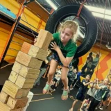 Girl moves through a suspended tire obstacle while stacking wooden blocks as campers watch during one of Mission Grit's team building summer camps for kids.