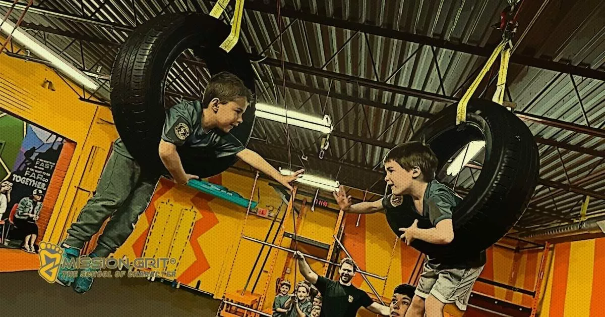 Two boys reach toward each other from hanging tire obstacles while teammates and coaches watch below.